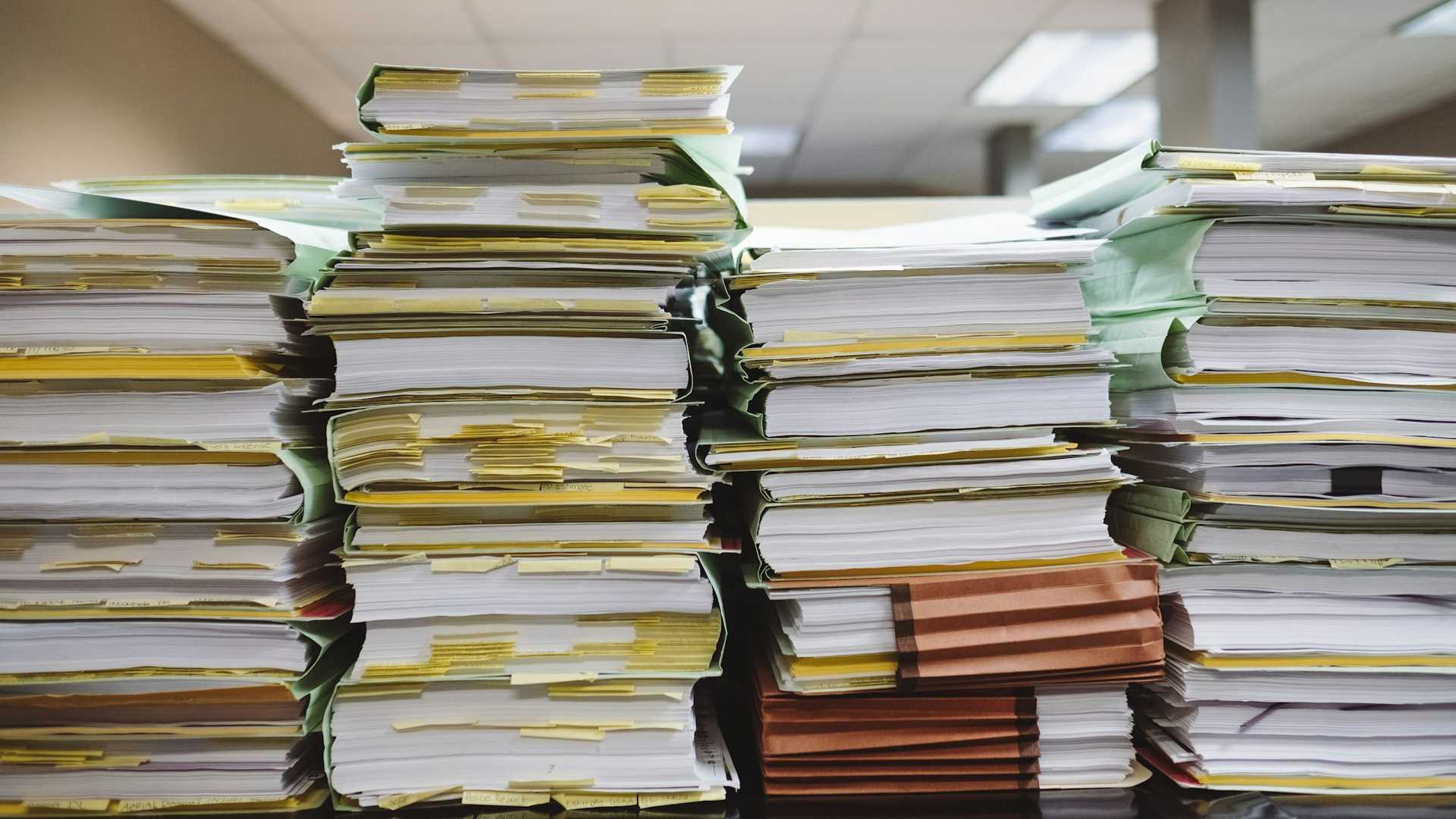Pilhas de processos, papéis e pastas de arquivo empilhados sobre uma mesa
